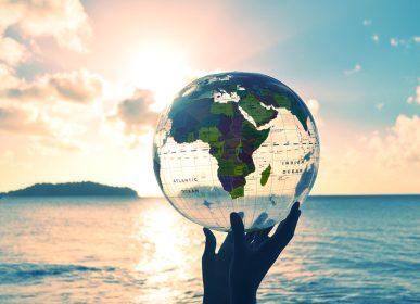 a transparent globe showing the map of Africa is held in silhouetted hands and raised against a bright sunset and beach horizon. A perfect concept about global environmental issues such as global warming and eco friendly and renewable energy themes
